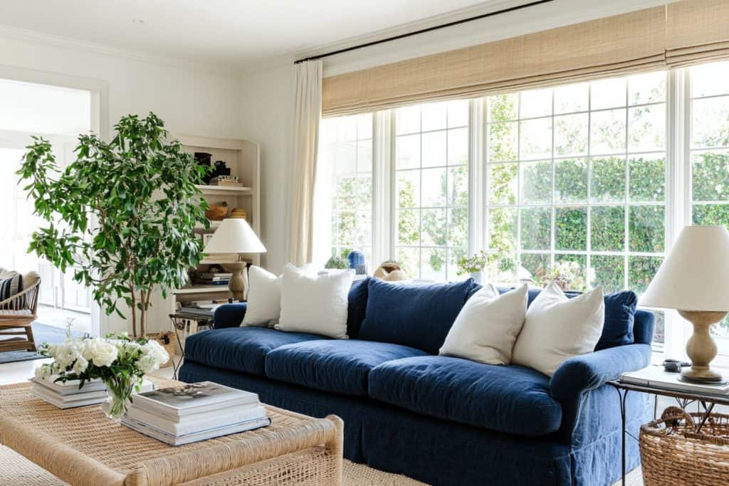 A classic living room with a blue velvet sofa, framed by two black-trimmed French doors and flanked by houseplants. A marble coffee table and framed botanical art complete the chic look.