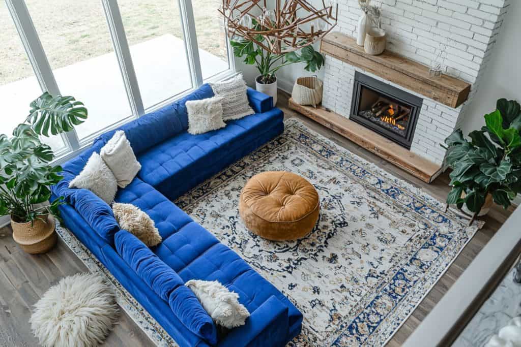 A bird's-eye view of a living room featuring a tufted blue sectional sofa and a circular brown ottoman. The room has large windows, a fireplace with wooden mantle, and indoor plants, all on a blue-pat