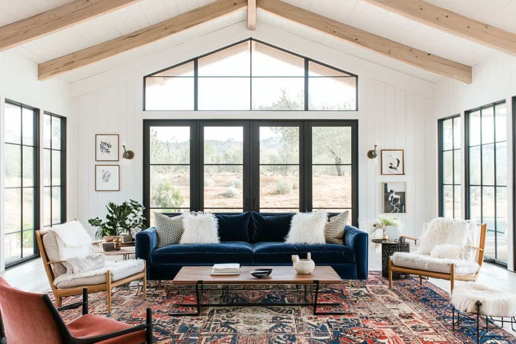 A living room with a rustic and airy feel, featuring a deep blue sofa against large black-framed windows. A vaulted ceiling with wooden beams and a colorful patterned rug enhance the open and warm atm