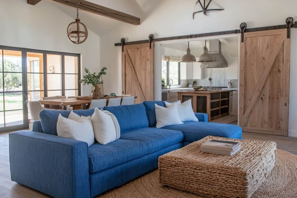 A rustic-themed living room with a modern blue sectional sofa and white throw pillows. The room includes sliding barn doors leading to the kitchen, a woven coffee table, and a dining area in the back.