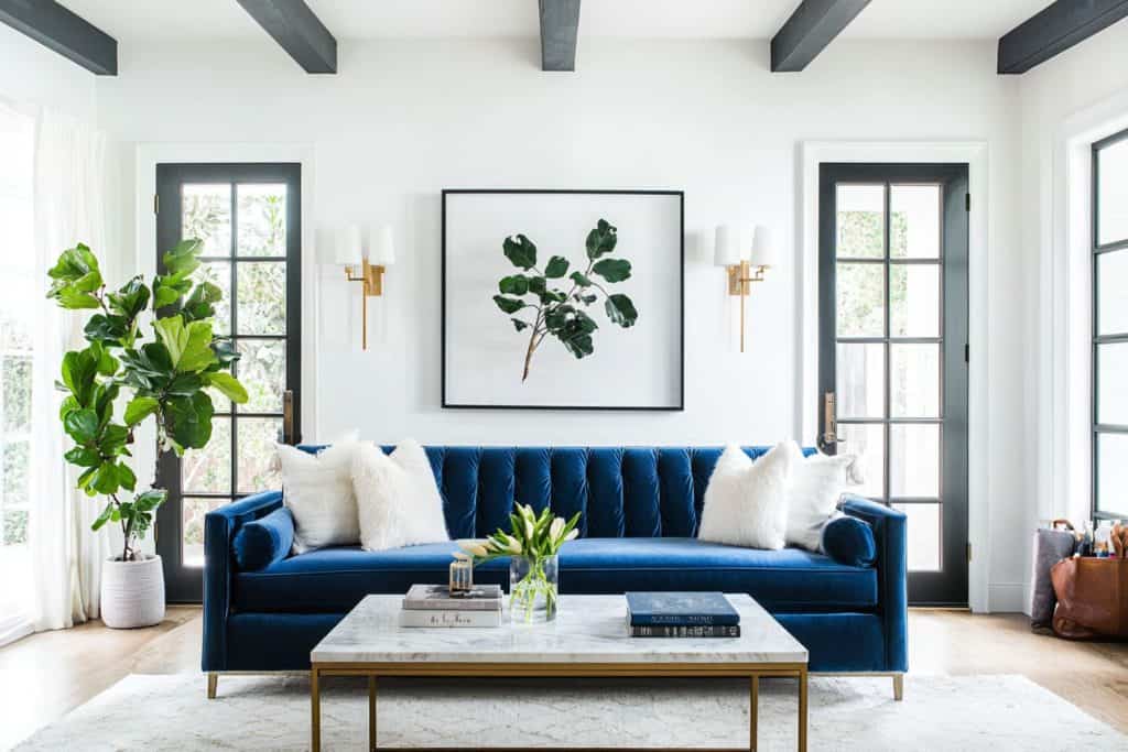 A minimalist living room featuring a deep blue velvet sofa against a white paneled wall. A marble-top side table with white flowers adds elegance to the otherwise simple and clean design.